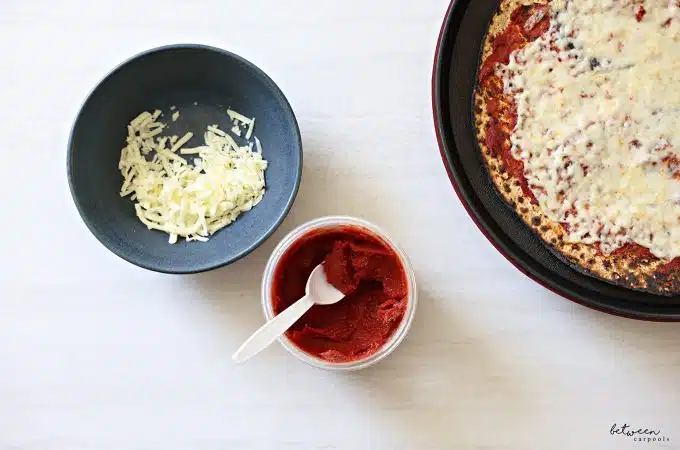 Top view of an open 16 oz twist top storage container with seasoned tomato paste and a plastic spoon labeled pizza sauce. One round Shmurah Matzah with a thin layer of seasoned tomato paste and cheese in a Betty Crocker. Bowl with some shredded cheese nearby.
