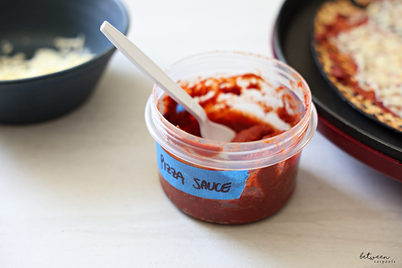 An open 16 oz twist top storage container with seasoned tomato paste and a plastic spoon labeled pizza sauce. One round Shmurah Matzah with a thin layer of seasoned tomato paste and cheese in a Betty Crocker. Bowl with some shredded cheese nearby.