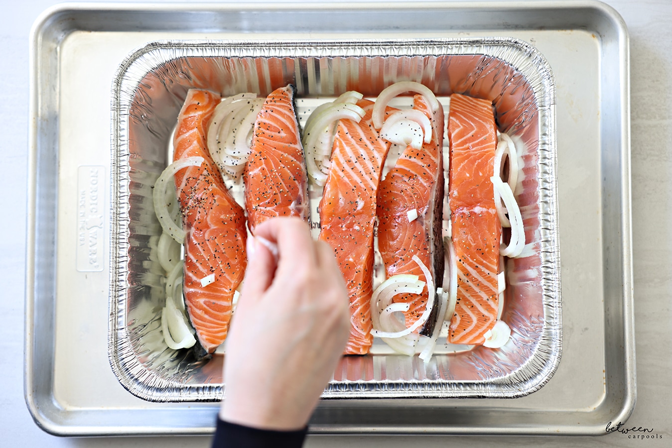 Slices of salmon, with salt pepper and thinly sliced onion in half moons, in a 9x13 disposable pan on a metal cookie sheet.