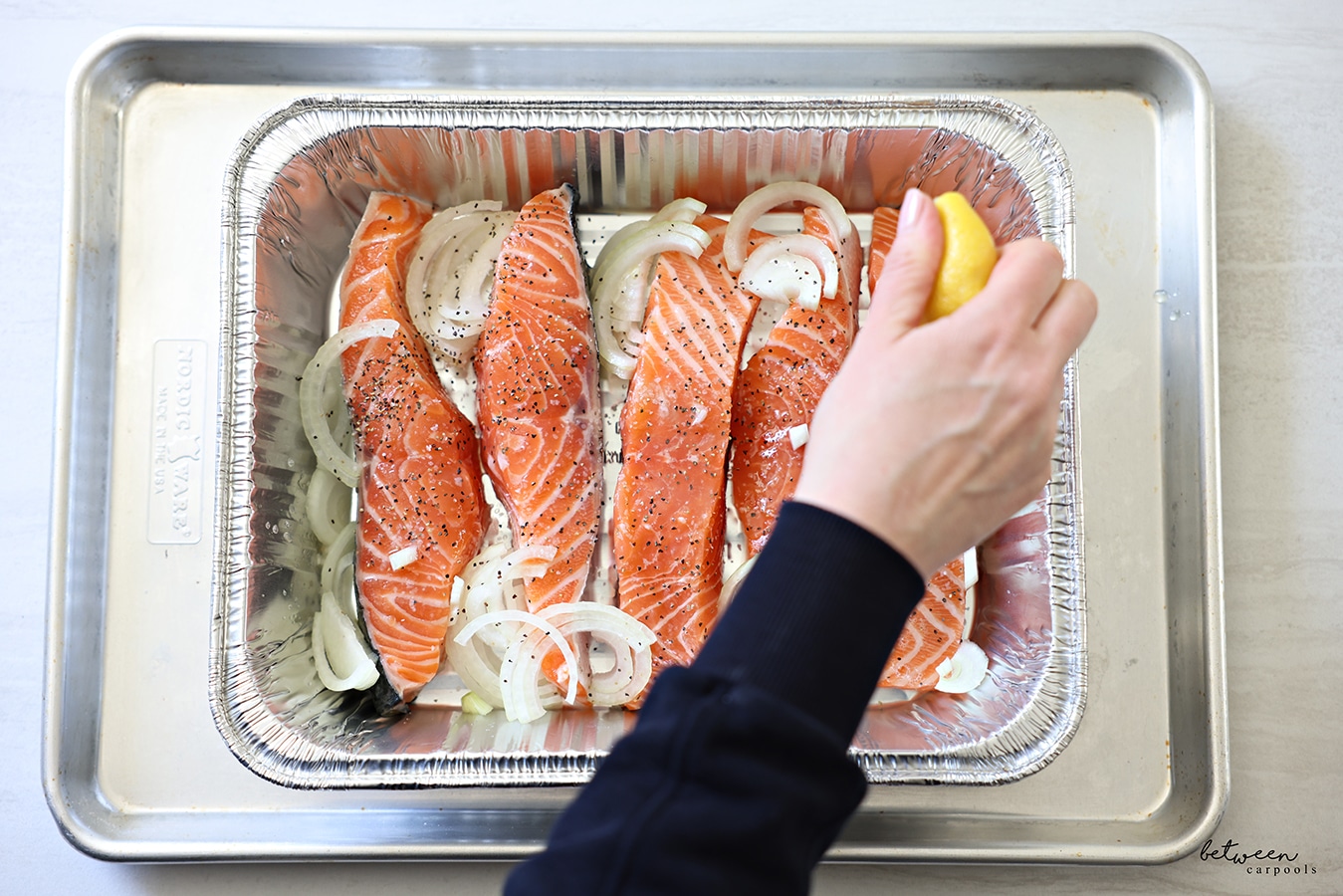Squeezing a lemon over slices of salmon, with salt pepper and thinly sliced onion in half moons, in a 9x13 disposable pan on a metal cookie sheet.