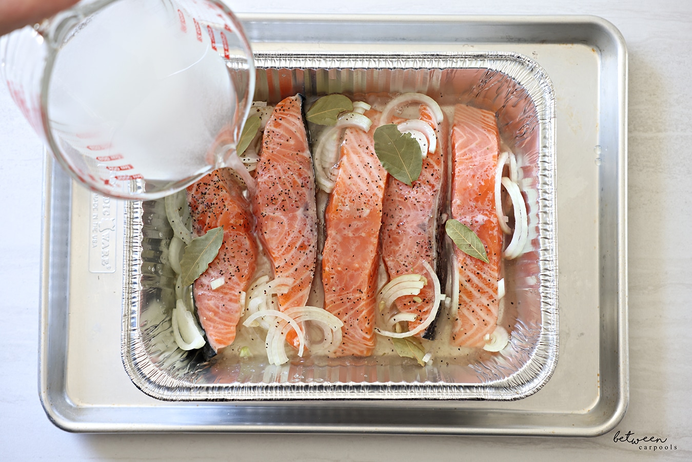 Pouring water, vinegar, and sugar over slices of salmon, with salt pepper thinly sliced onion in half moons and lemon, in a 9x13 disposable pan on a metal cookie sheet.