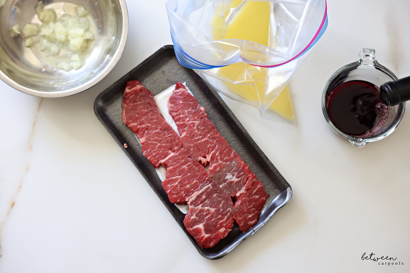 Grated onion in a metal mixing bowl, a package of strip steaks, orange juice in an open Ziploc bag, and pouring red wine into a glass measuring cup.