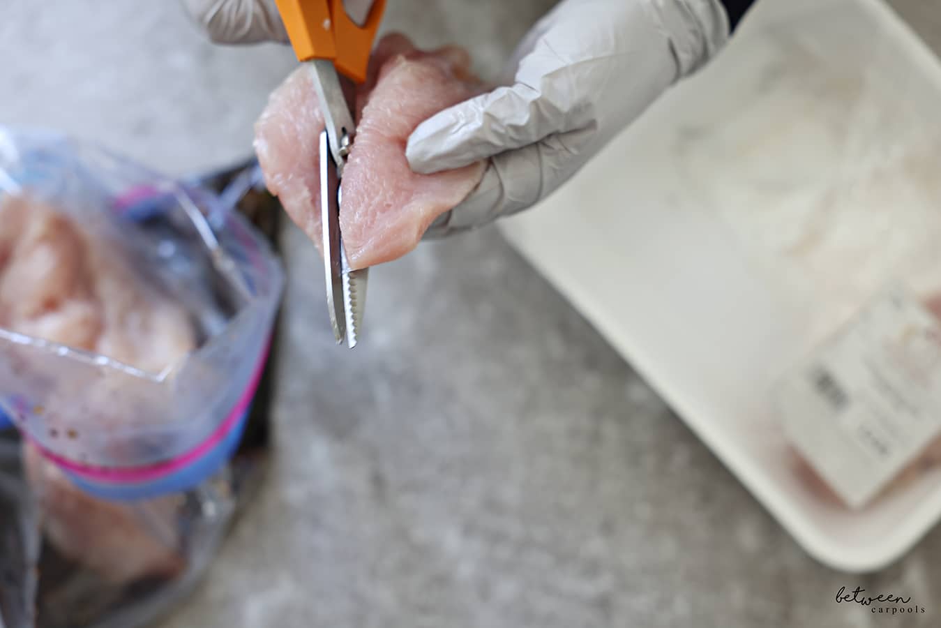 Cutting chicken breast with kitchen scissors and gloved hands. Resealable bag with chicken + marinade, and a package of chicken nearby.