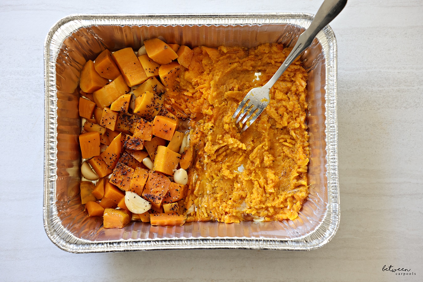 A 9x13 pan with baked sweet potato chunks and whole garlic cloves, in oil + salt and pepper, half mashed with a fork.