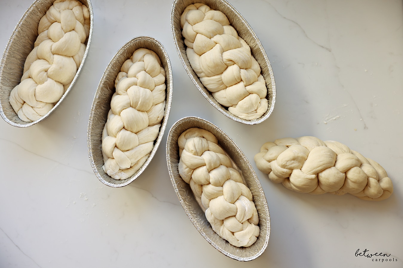 4 braided challah in foil challah pans. 1 braided challah on the kitchen counter.