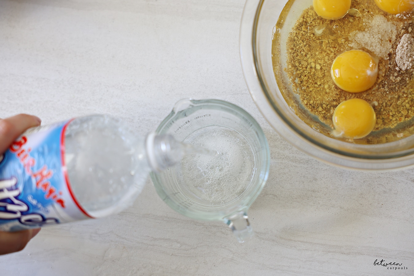 Pouring ¼ cup Be'er Mayim Seltzer into a glass measuring cup. Glass mixing bowl with Matzah Ball ingredients (eggs, oil, and matzah meal) nearby.
