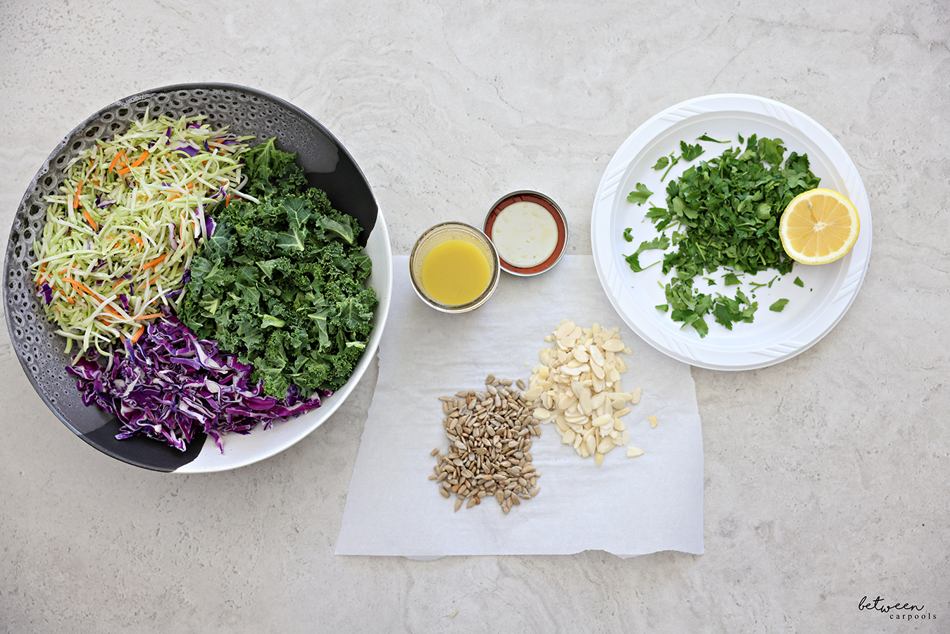 Broccoli slaw, finely chopped kale and shredded red cabbage in a large salad bowl. Sunflower seeds and sliced almonds on a piece of parchment. Lemon vinaigrette in an open Mason jar and a plastic plate with chopped parsley and a half a lemon.