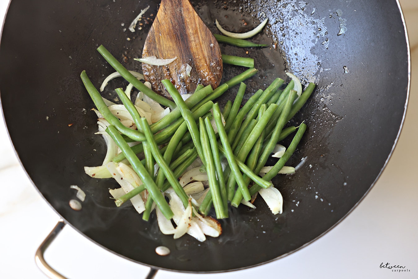 Green beans added to a Frenched onion and minced garlic sautéing in a wok with a wooden spoon.