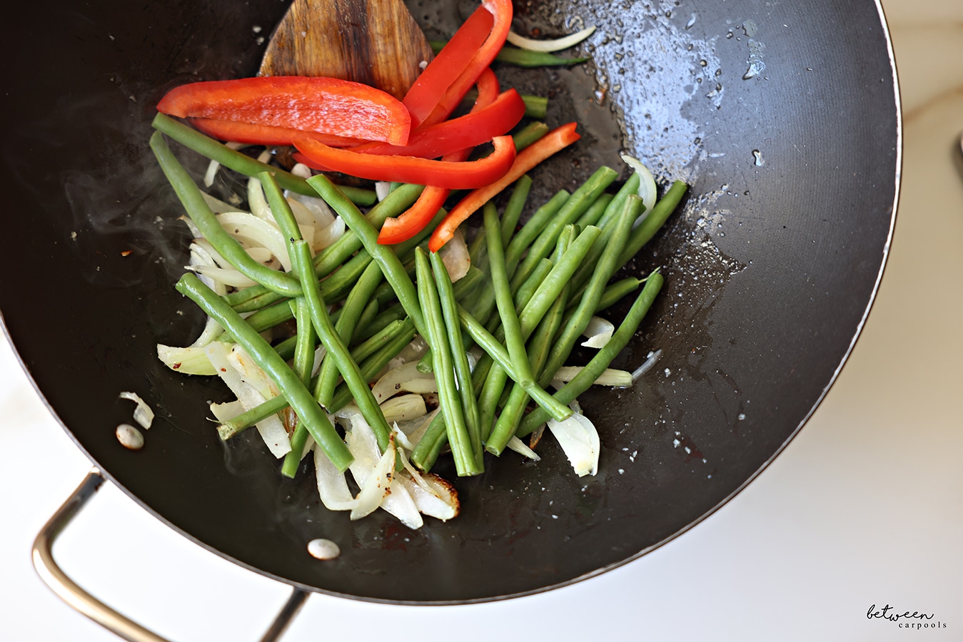Green beans and sliced red pepper added to a Frenched onion and minced garlic sautéing in a wok with a wooden spoon.