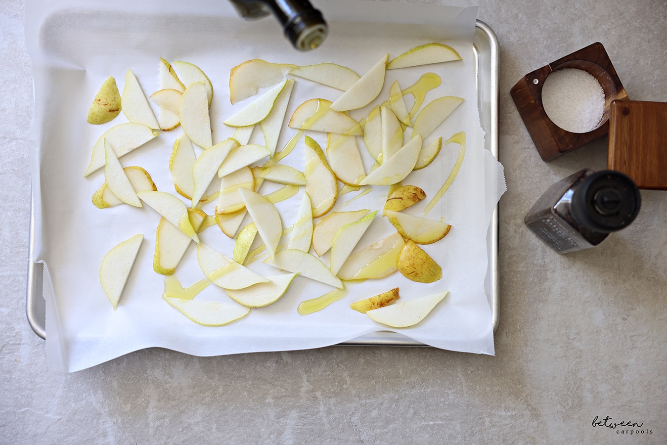 Drizzling olive oil over pear slices on a parchment lined baking sheet. Salt cellar and black pepper grinder nearby.