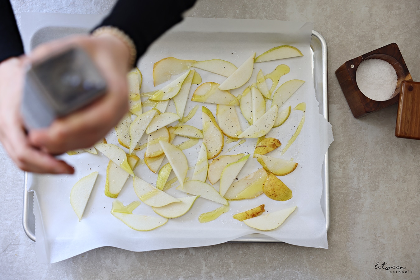 Grinding black pepper over pear slices drizzled with olive oil on a parchment lined baking sheet. Salt cellar nearby.