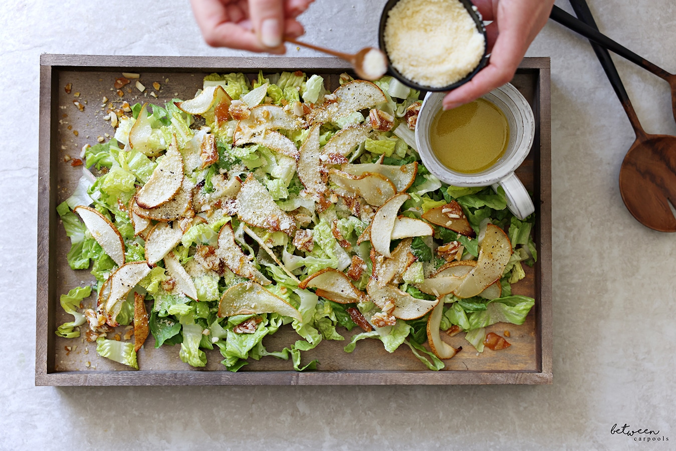 Sprinkling Parmesan over Romaine lettuce, roasted pears, and crushed praline, on a wooden board. Creamer with salad dressing on the board.