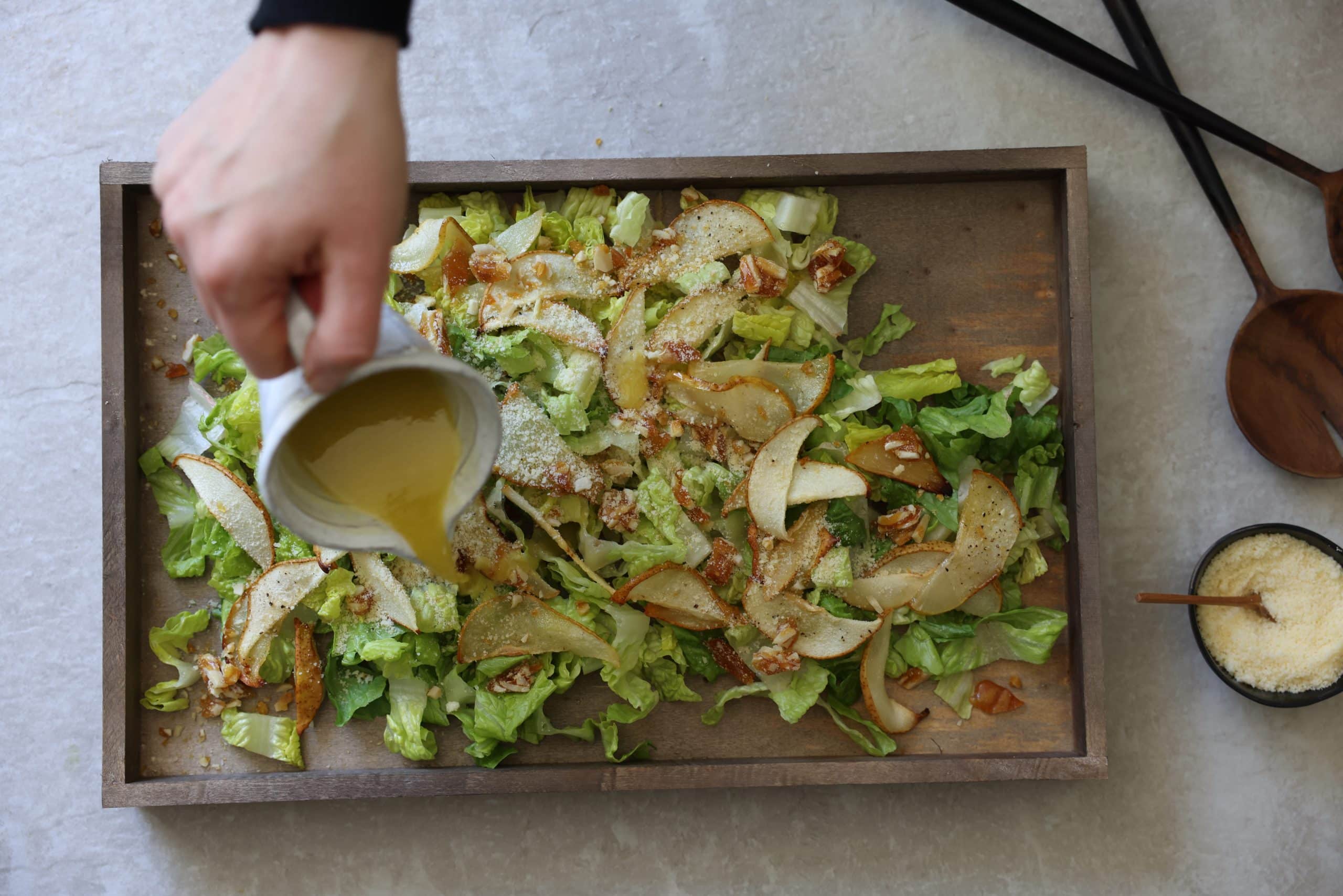 Pouring salad dressing over Romaine lettuce, roasted pears, crushed praline, and Parmesan cheese. on a wooden board.