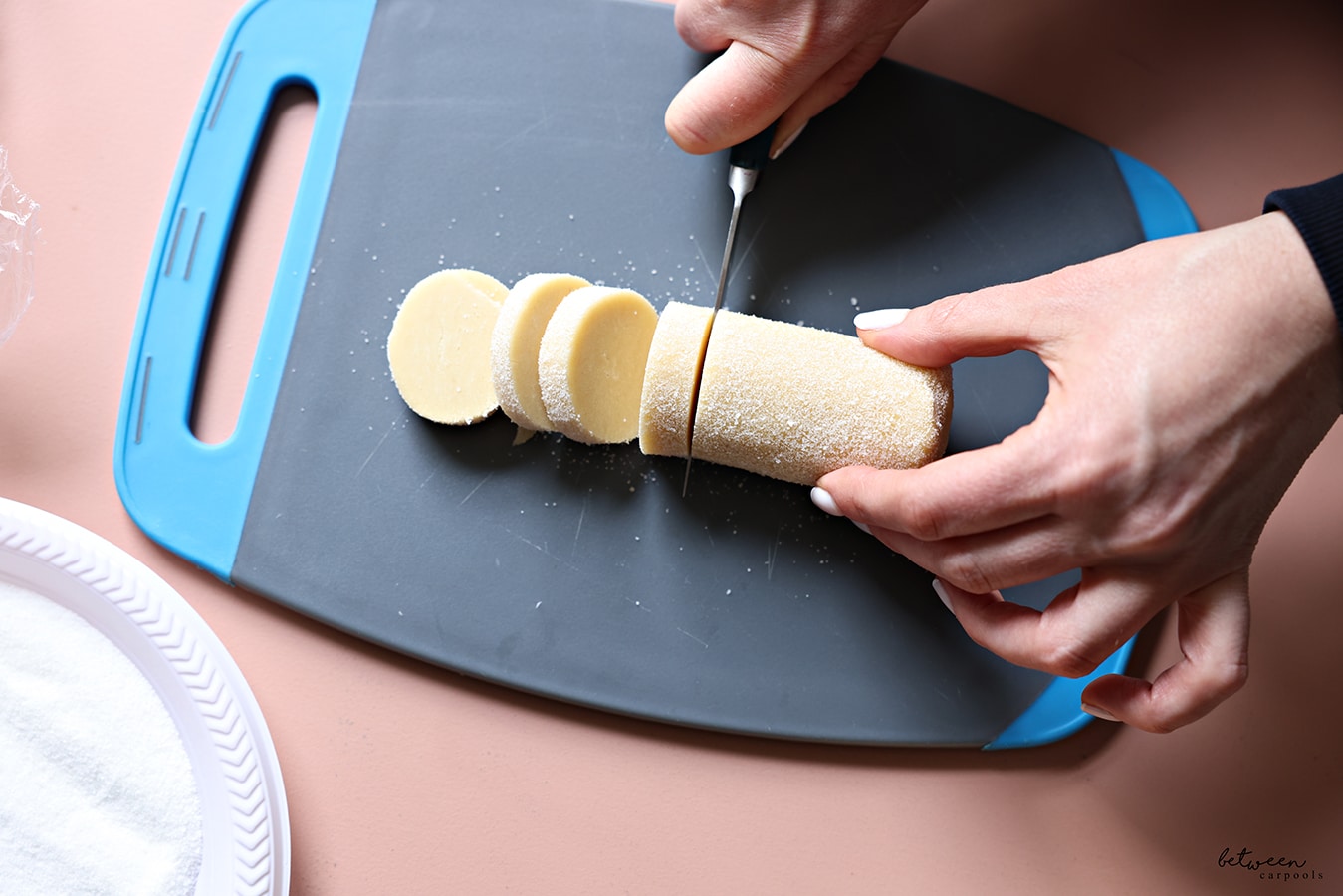 Cutting one log of French Butter Cookie dough rolled in sugar into slices on a cutting board. A plastic plate with sugar nearby.