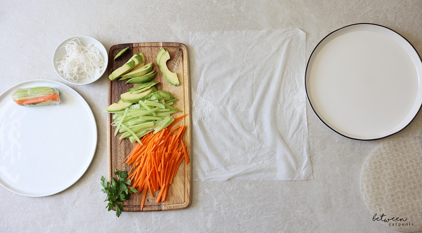 R to L: rice paper wrappers, a shallow plate with warm water, a damp paper towel, a wooden cutting board with cucumbers and carrots cut into matchsticks, sliced avocado and fresh parsley, a bowl of cooked rice noodles, and a plate with one finished spring roll.