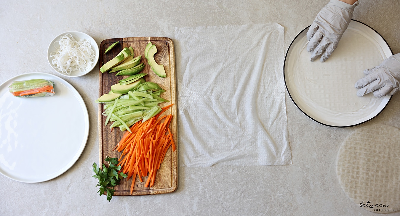 Dipping one sheet of rice paper into warm water with gloved hands. R to L: rice paper wrappers, a shallow plate with warm water, a damp paper towel, a wooden cutting board with cucumbers and carrots cut into matchsticks, sliced avocado and fresh parsley, a bowl of cooked rice noodles, and a plate with one finished spring roll.