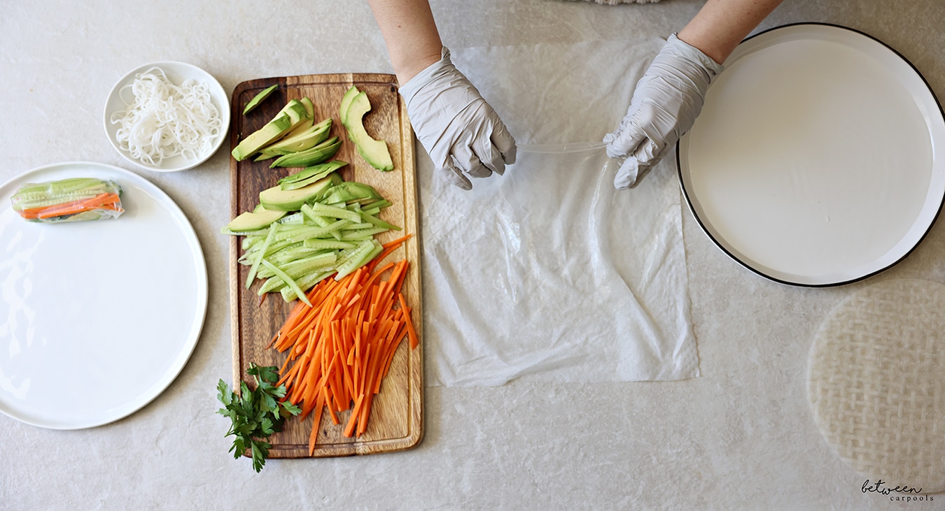 Transferring one sheet of rice paper dipped into warm water to a damp paper towel. R to L: rice paper wrappers, a shallow plate with warm water, a damp paper towel, a wooden cutting board with cucumbers and carrots cut into matchsticks, sliced avocado and fresh parsley, a bowl of cooked rice noodles, and a plate with one finished spring roll.
