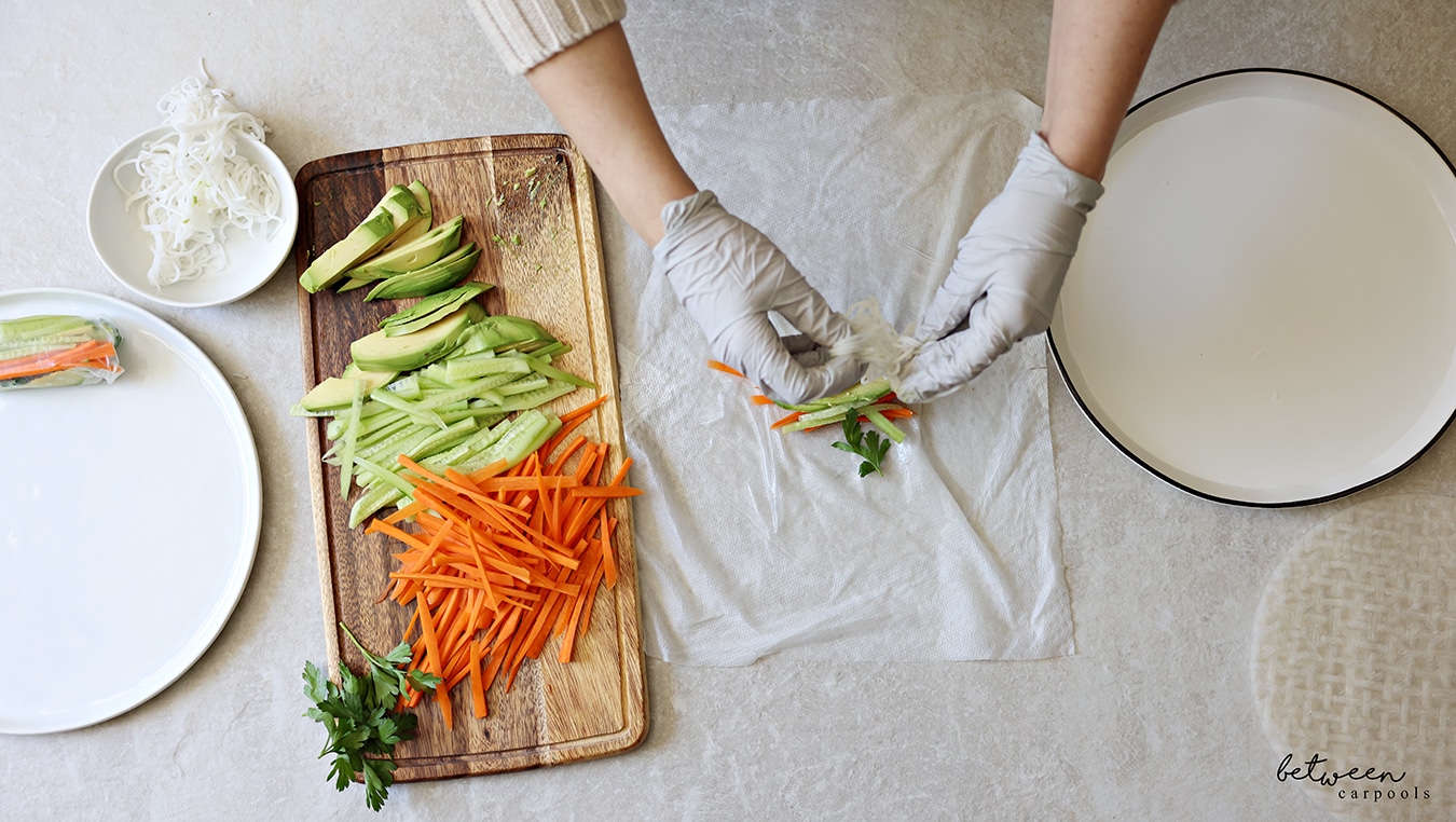 Adding rice noodles to one sheet of rice paper, dipped into warm water, on a damp paper towel. R to L: rice paper wrappers, a shallow plate with warm water, a damp paper towel, a wooden cutting board with cucumbers and carrots cut into matchsticks, sliced avocado and fresh parsley, a bowl of cooked rice noodles, and a plate with one finished spring roll.