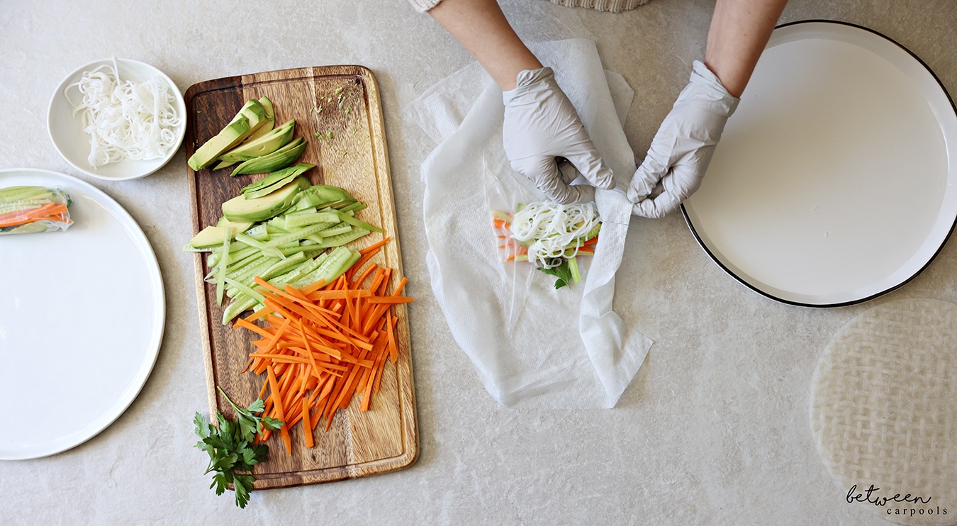 Folding in the sides of a rice paper with filling. R to L: rice paper wrappers, a shallow plate with warm water, a damp paper towel, a wooden cutting board with cucumbers and carrots cut into matchsticks, sliced avocado and fresh parsley, a bowl of cooked rice noodles, and a plate with one finished spring roll.
