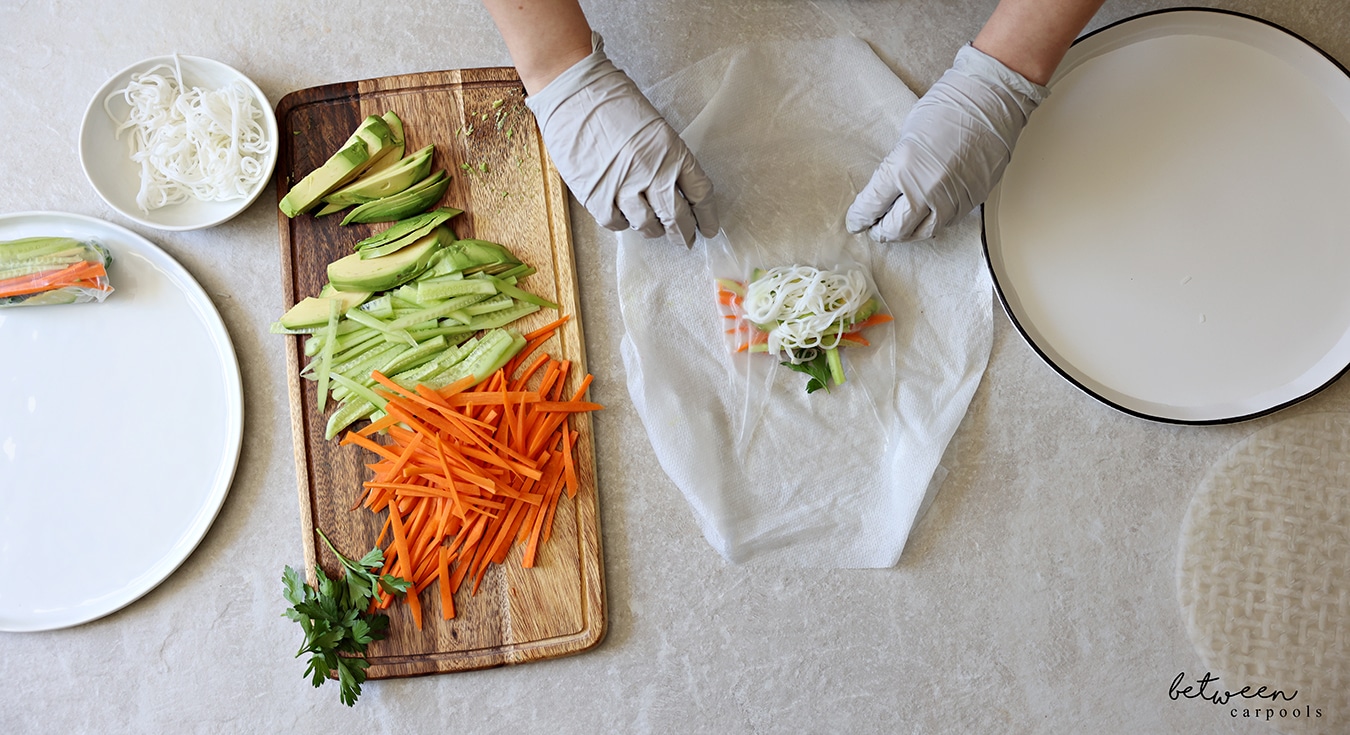 Folding the bottom of a rice paper over the filling. R to L: rice paper wrappers, a shallow plate with warm water, a damp paper towel, a wooden cutting board with cucumbers and carrots cut into matchsticks, sliced avocado and fresh parsley, a bowl of cooked rice noodles, and a plate with one finished spring roll.