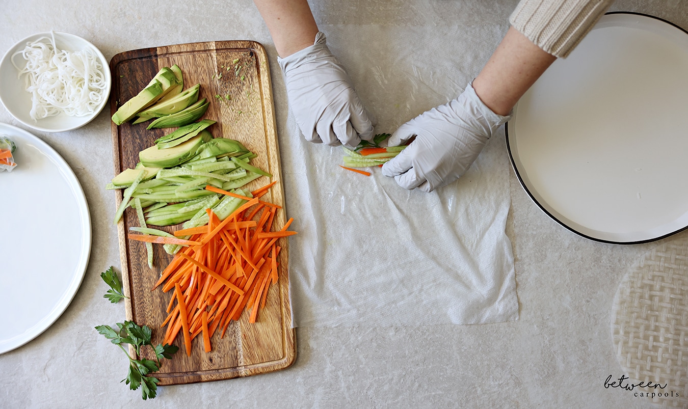 Adding filling to one sheet of rice paper, dipped into warm water, on a damp paper towel. R to L: rice paper wrappers, a shallow plate with warm water, a damp paper towel, a wooden cutting board with cucumbers and carrots cut into matchsticks, sliced avocado and fresh parsley, a bowl of cooked rice noodles, and a plate with one finished spring roll.