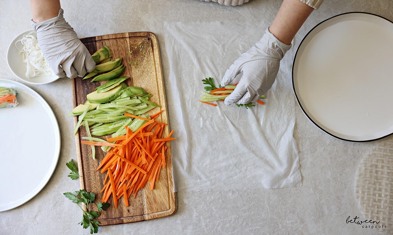 Adding avocado filling to one sheet of rice paper, dipped into warm water, on a damp paper towel. R to L: rice paper wrappers, a shallow plate with warm water, a damp paper towel, a wooden cutting board with cucumbers and carrots cut into matchsticks, sliced avocado and fresh parsley, a bowl of cooked rice noodles, and a plate with one finished spring roll.