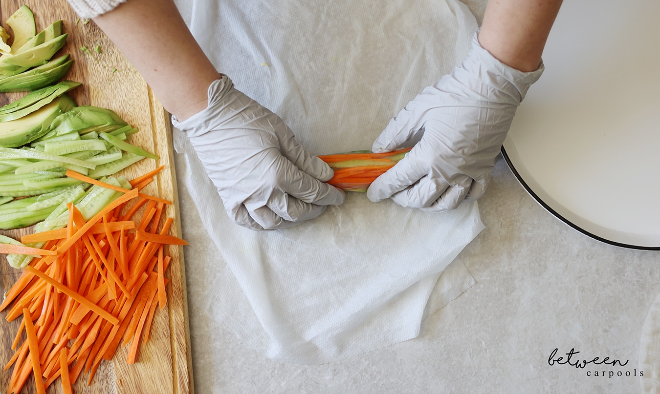 Rolling a filled rice paper tightly, on a damp paper towel, with gloved hands.