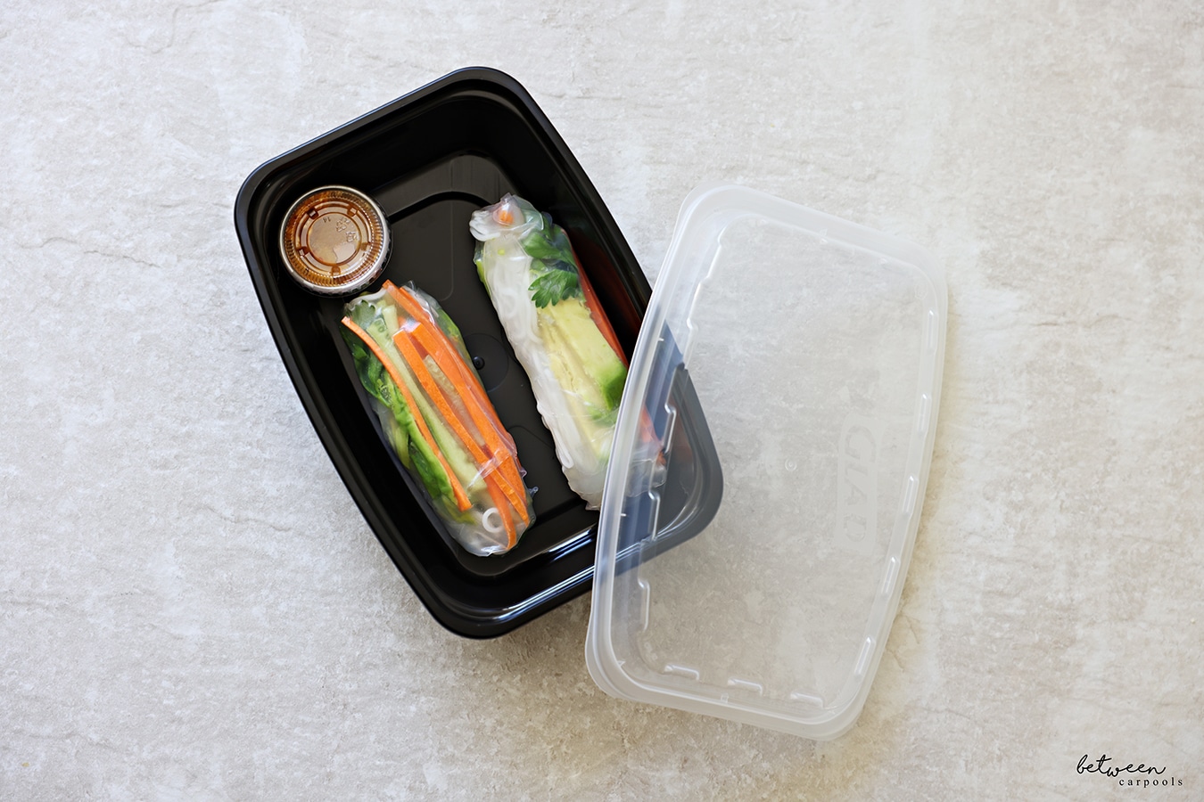 Two spring rolls and a condiment cup with peanut dipping sauce in a meal prep plastic food container.
