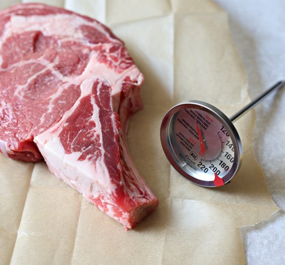 An Escali Stainless Steel Oven Safe Meat Thermometer near a bone-in ribeye on a piece of natural parchment paper.