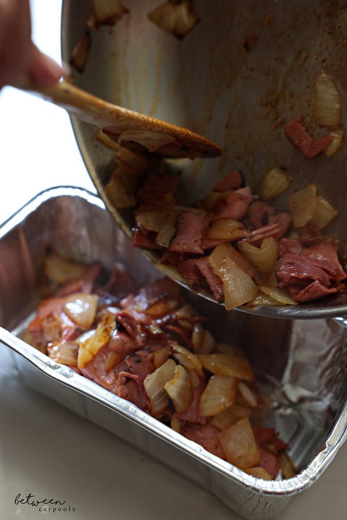 Transferring sautéed onion chunks and deli pastrami cubes with BBQ sauce from a sauté pan into an aluminum loaf pan with a wooden paddle.