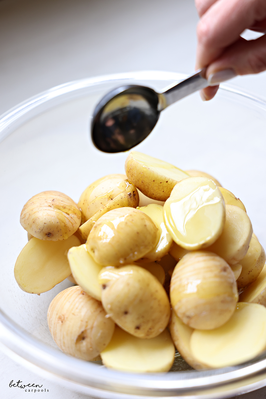 Adding olive oil to a glass bowl with hasselback baby potato halves.