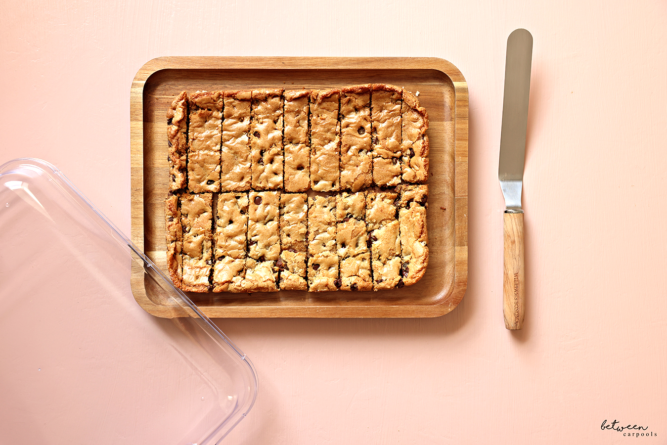 Between Carpools 9×13 Life: Chocolate Chip Sticks on a bamboo tray. Lid and Williams Sonoma Olivewood Straight Icing Spatula nearby.