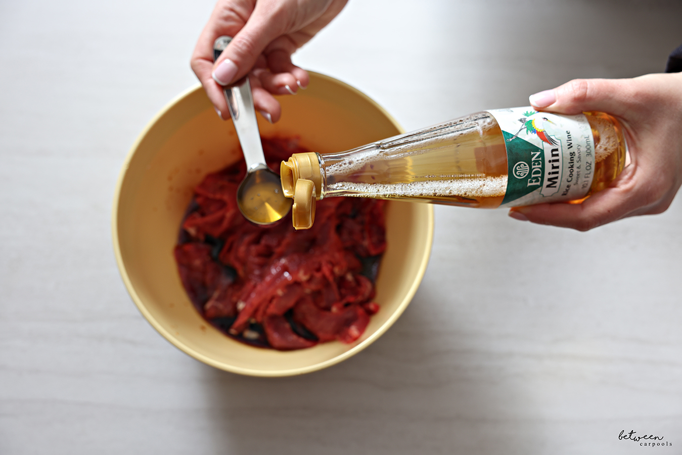 Measuring mirin over a bowl with thinly sliced steak and marinade.