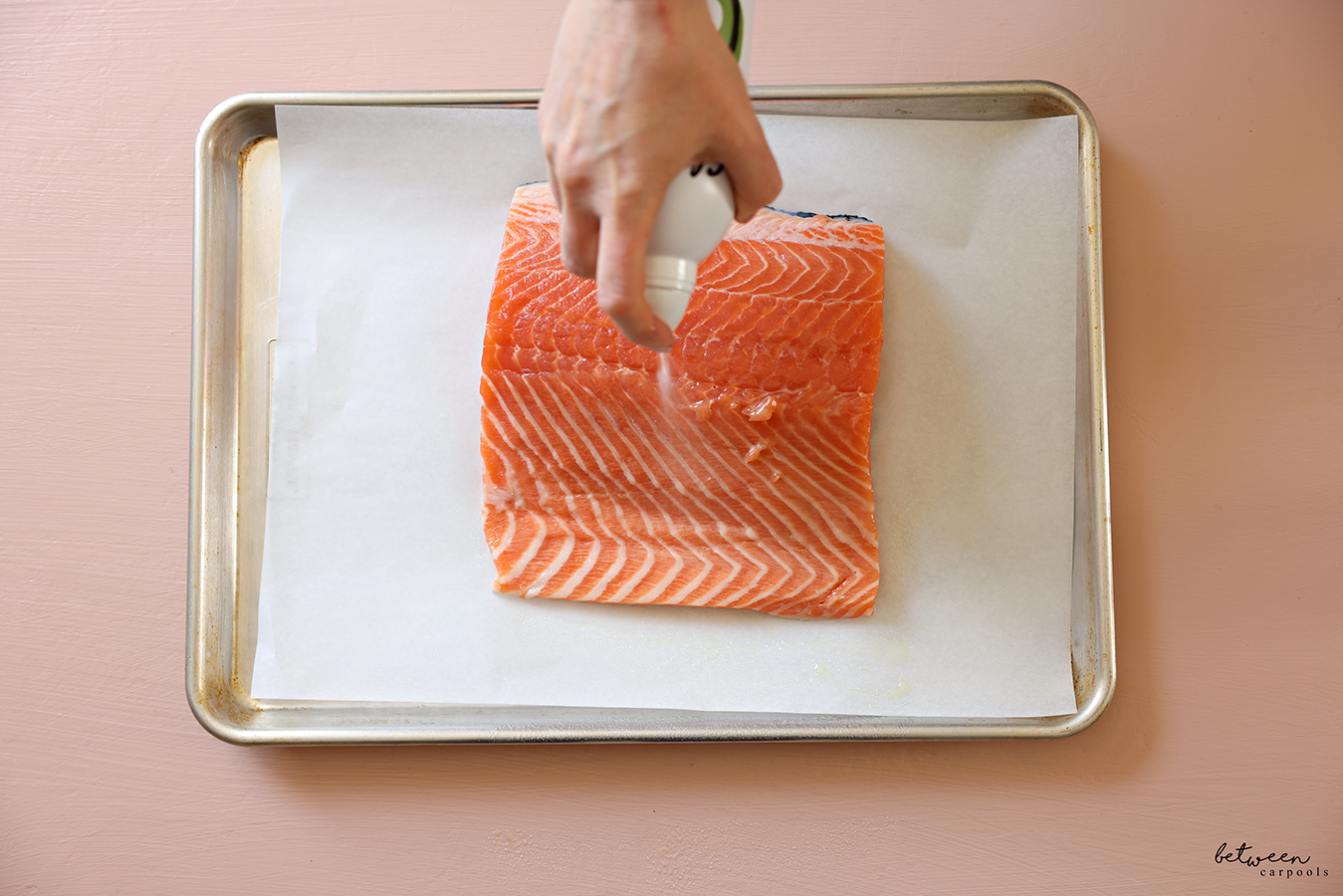 Spraying a piece of salmon on a parchment lined baking sheet with avocado oil.