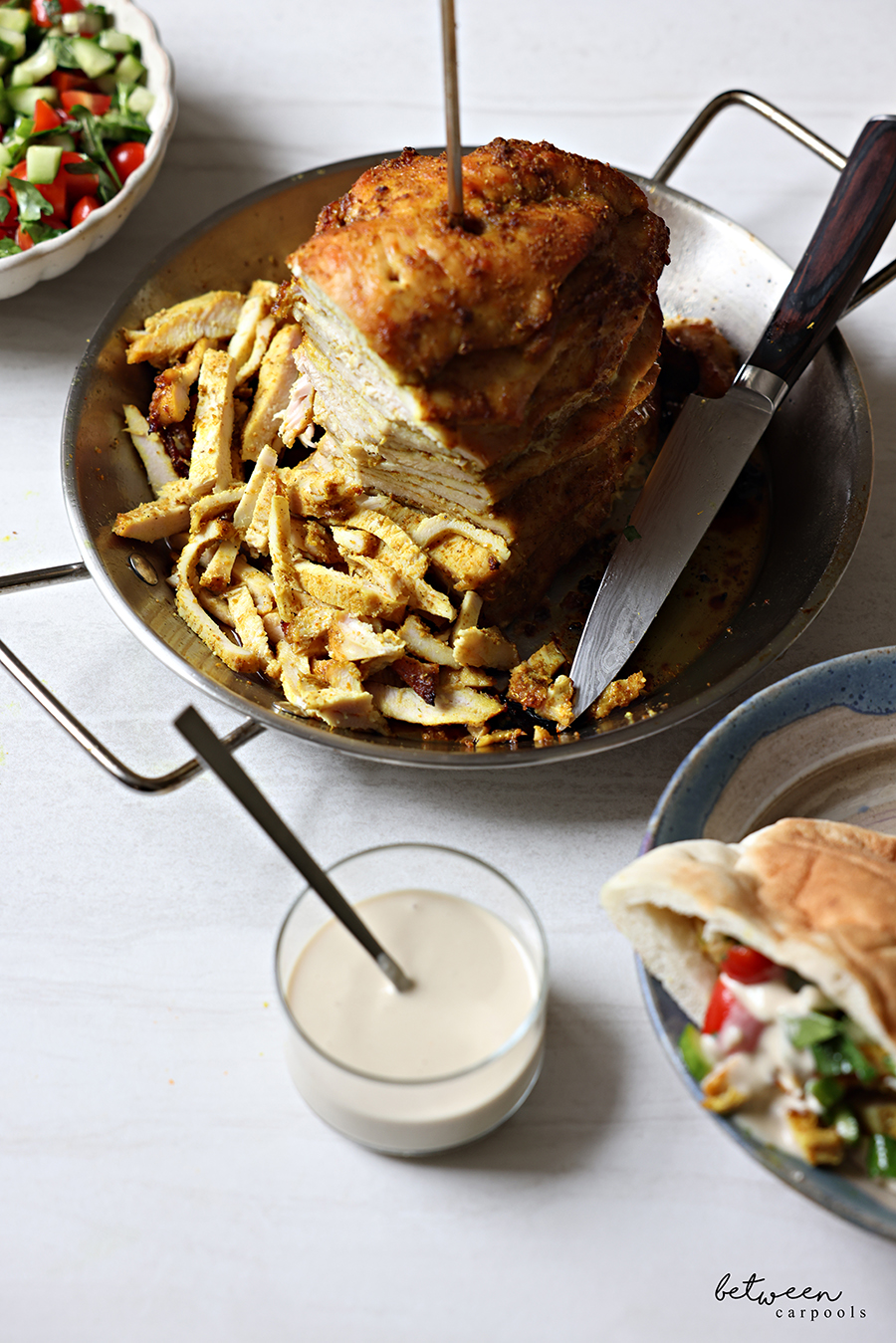 Sliced shawarma chicken off the skewer in an Al Pastor Vertical Skewer, a bowl of salad, a glass of techina, and a pita with those components plated.