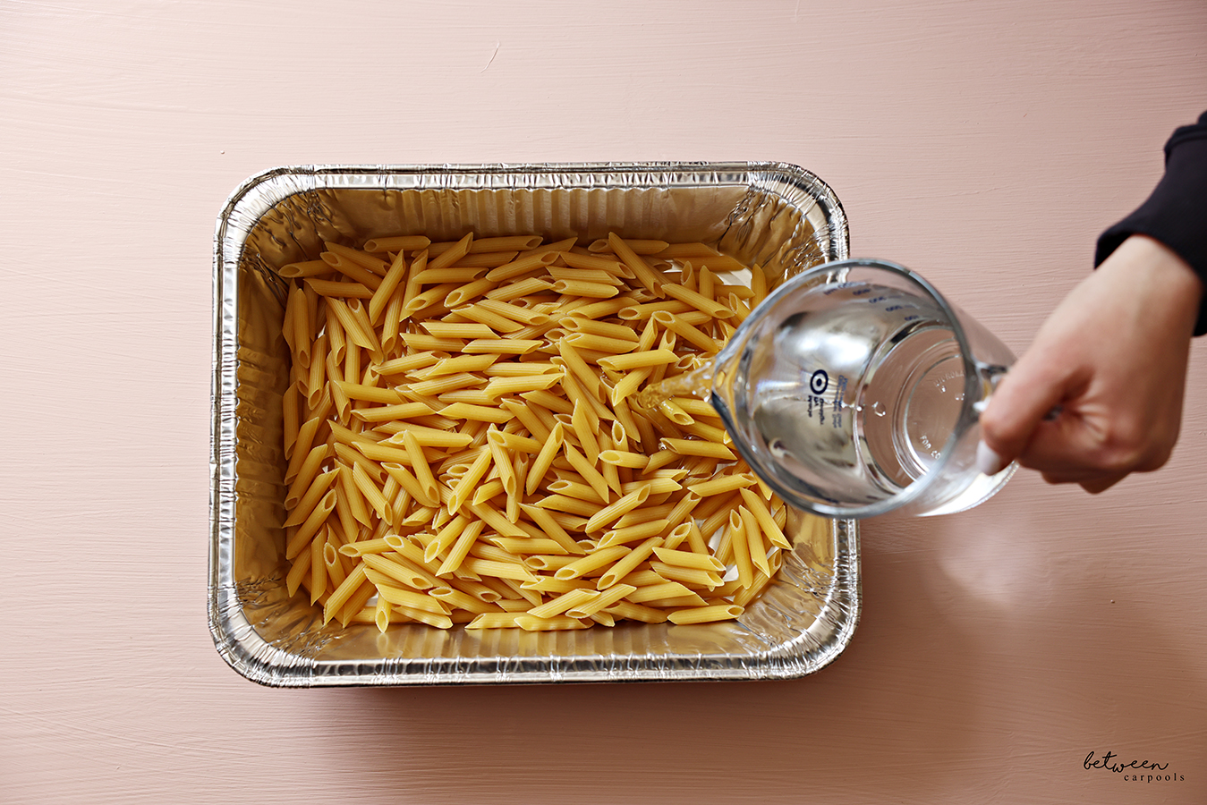 Adding water to penne pasta in a 9x13-inch aluminum foil pan.