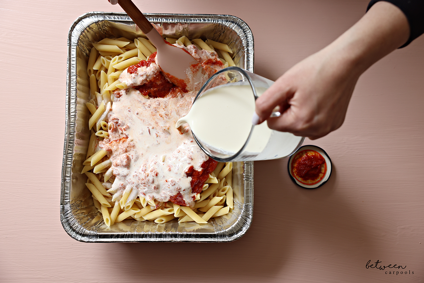 Pouring heavy cream over marinara sauce and pasta in a 9x13-inch aluminum foil pan.