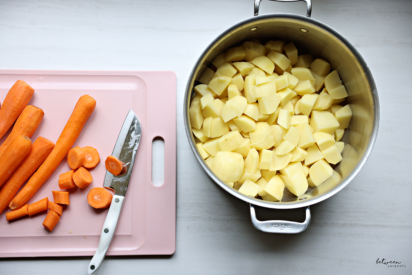 Top view of peeled and cubed potatoes in a large pot and peeled carrots on a pink cutting board.