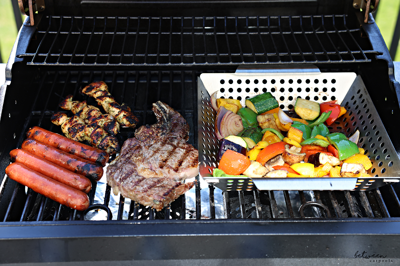Hot dogs, chicken, steak, and vegetables in a grill basket, on the grill.