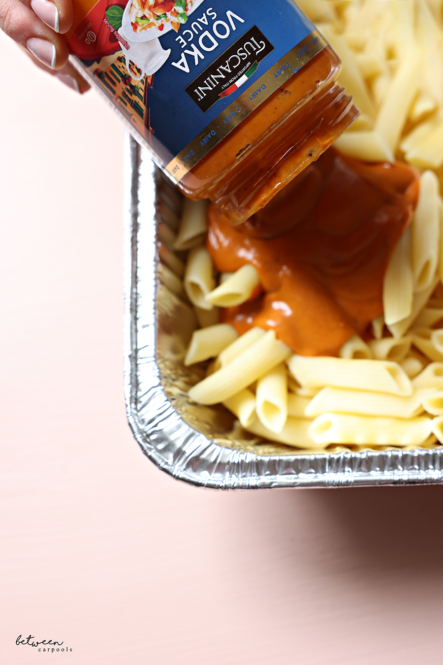 Pouring Tuscanini Vodka Sauce over cooked penne pasta in a 9x13-inch aluminum foil pan.