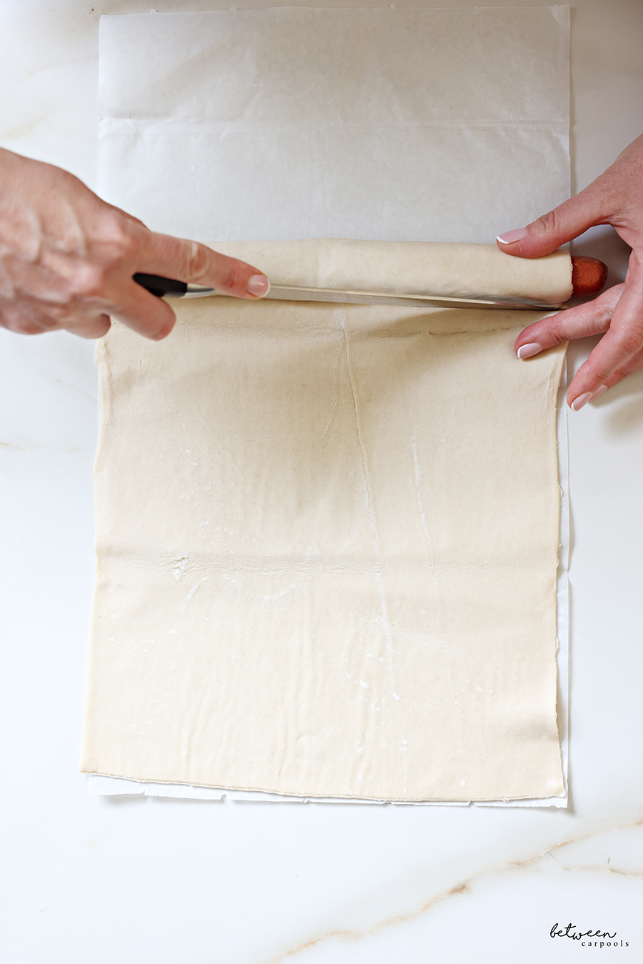 Cutting wrapped hot dogs in puff pastry from a sheet of pastry to place onto a piece of parchment paper.
