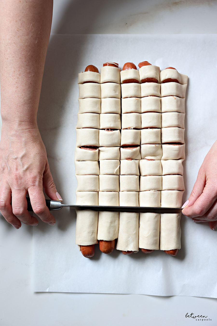 Cutting across hot dogs wrapped in puff pastry lined up [horizontally] next to each other.