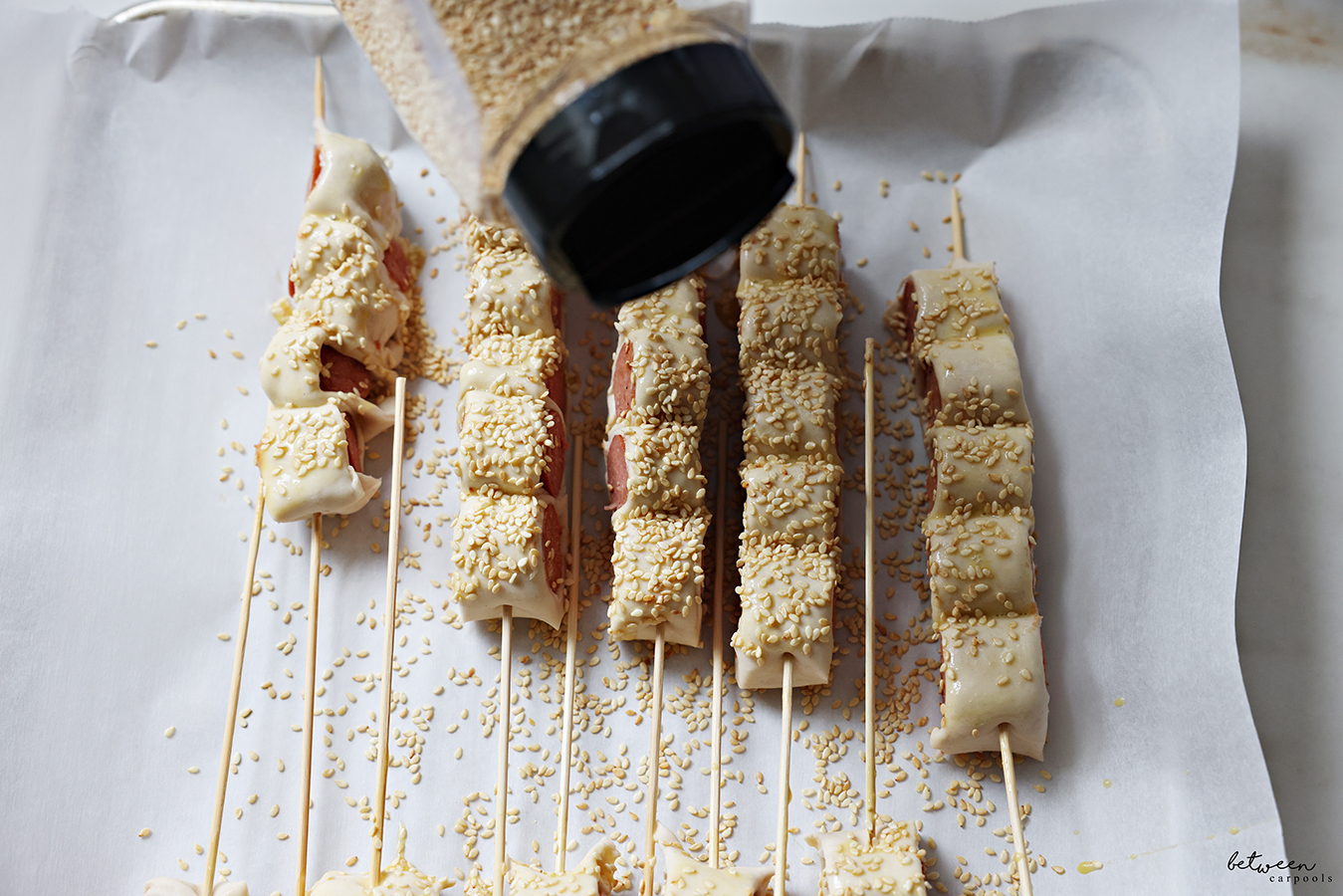 Generously sprinkling Puff Pastry Hot Dogs on a Stick with sesame seeds.