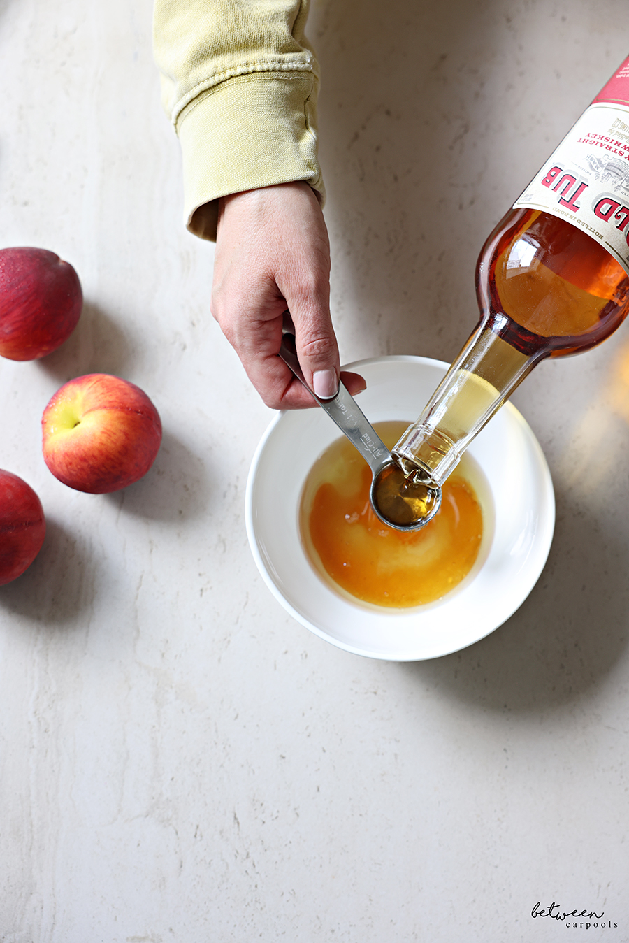 Adding bourbon to a small bowl with honey. Peaches nearby.