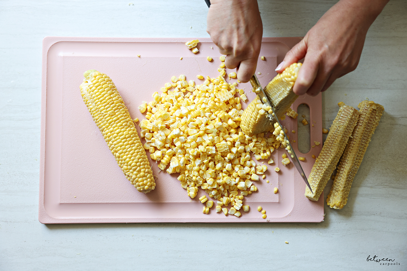 Slicing kernels off the cob of four ears of fresh corn, on a pink plastic cutting board.
