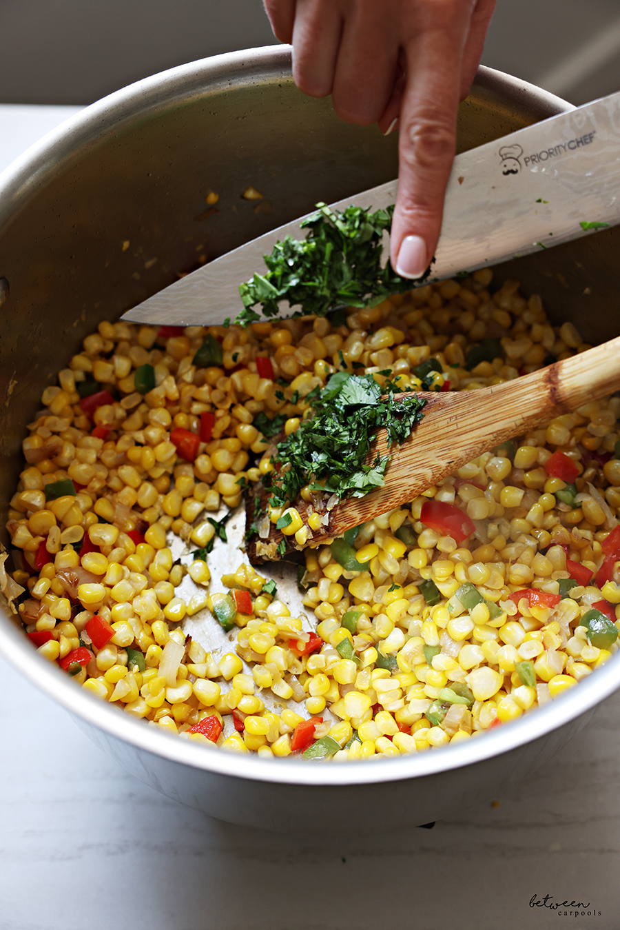 Adding chopped fresh cilantro to a sauté pan with shallot, jalapẽno, red bell pepper and corn kernels.