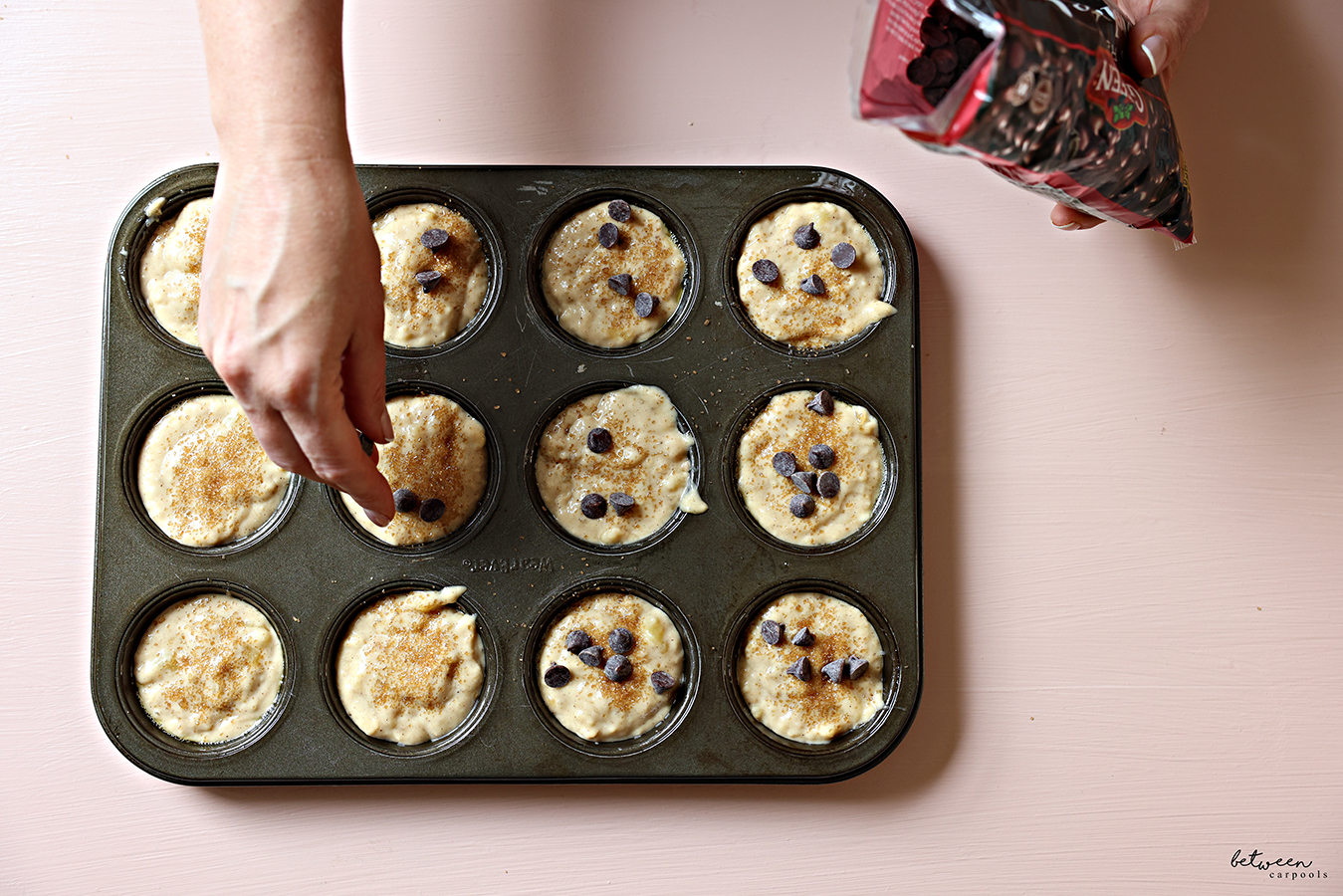Adding chocolate chips to banana muffins topped with turbinado sugar in a standard 12 cup muffin pan.