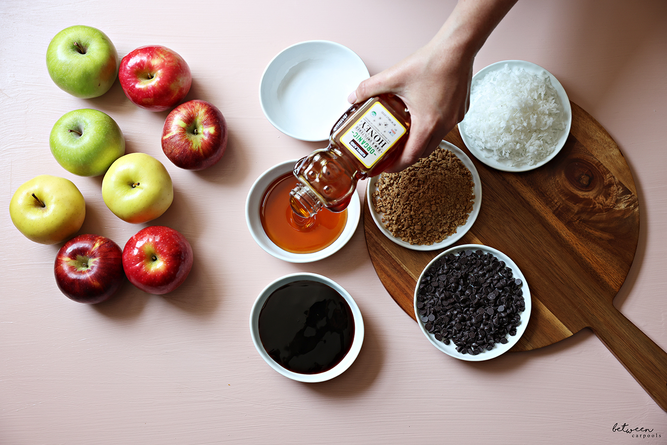 Caramel sauce, mini chocolate chips, Lotus Crunchies and shredded coconut flakes in shallow bowls on a wooden board. Pouring honey into another bowl. A variety of apples nearby.