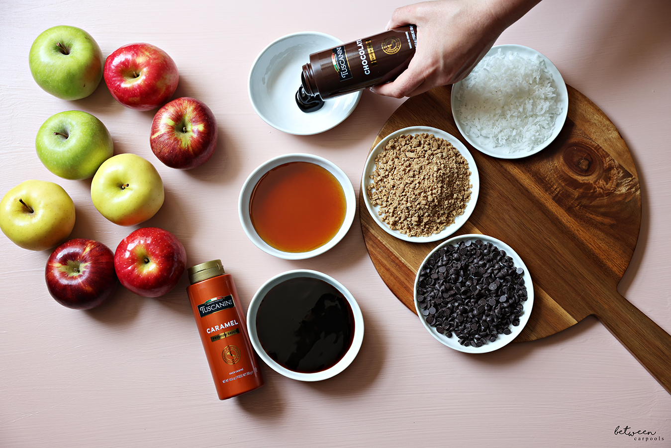 Honey, caramel sauce, mini chocolate chips, Lotus Crunchies and shredded coconut flakes in shallow bowls on a wooden board. Pouring Tuscanini Chocolate into another bowl. A variety of apples nearby.