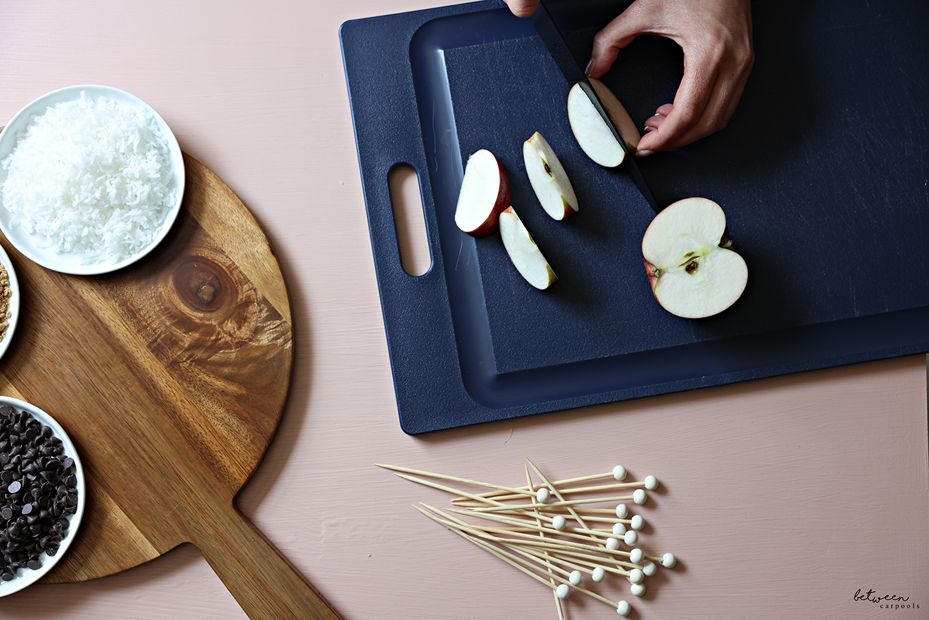Cutting half an apple into spears on a navy plastic cutting board. Skewers and toppings in shallow bowls nearby.